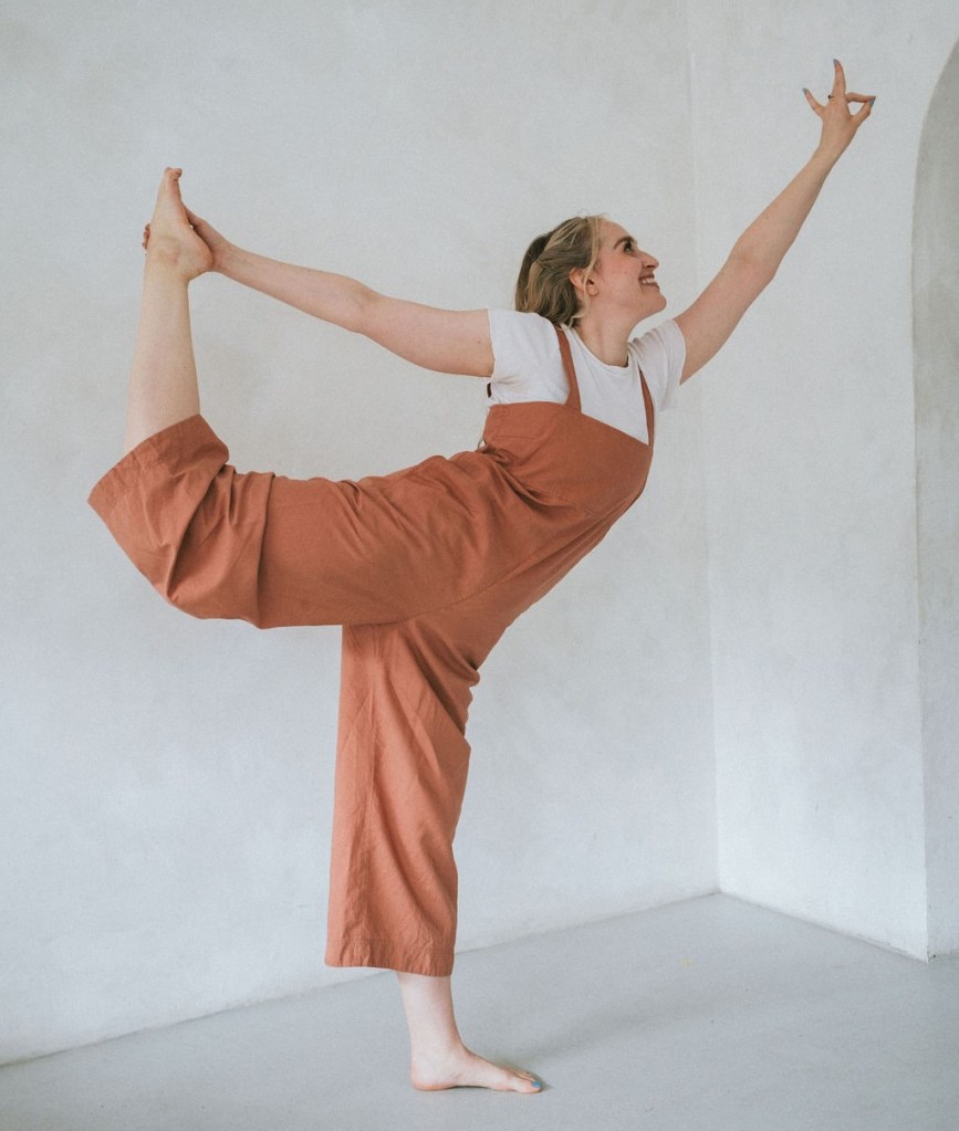 Yoga teacher in a terracotta jumpsuit performing dancer pose.