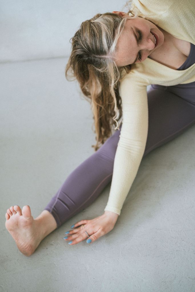 Yoga teacher in lilac leggings and a yellow cardigan performing a seated side bend