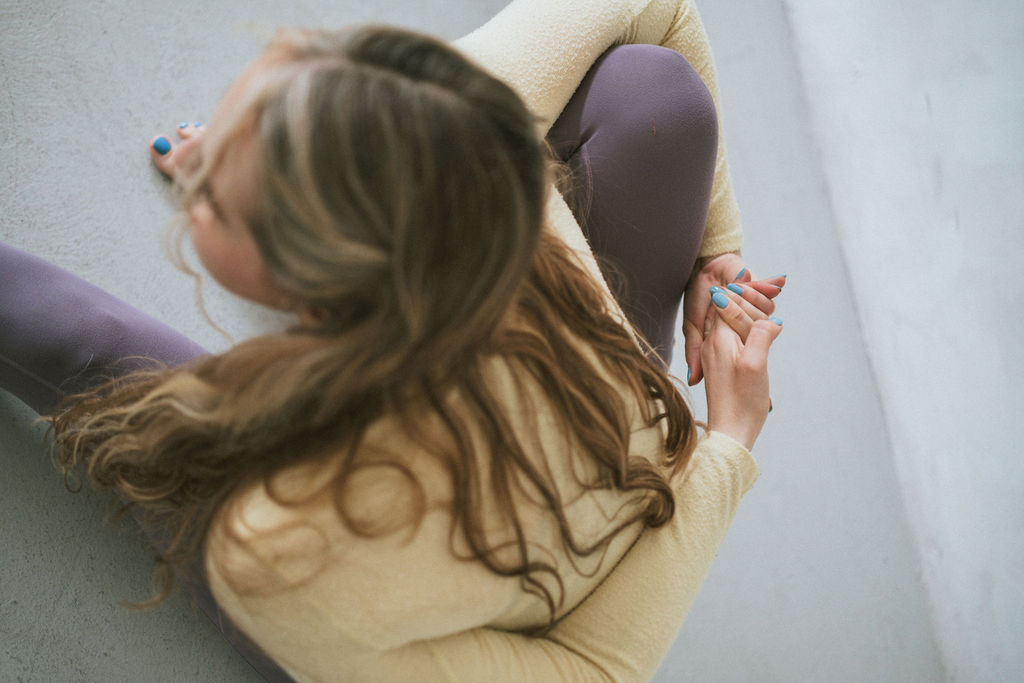 Yoga teacher in lilac leggings and lemon yellow cardigan performing Marciyasana A pose, a seated bound forward fold